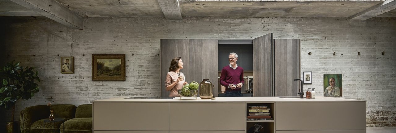Banner showing a modern industrial kitchen with an exposed concrete ceiling and a skylight, illuminating a large neutral-toned kitchen island. Two people interact casually: a woman with glasses and a gray-haired man. In the background, a white brick wall with paintings and, on the left, a green velvet sofa and a decorative plant.